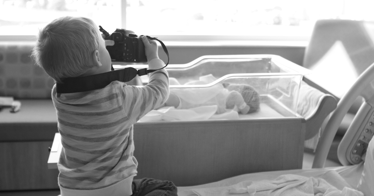 Black and white image of a young child using a digital camera to photograph a newborn in a bassinet