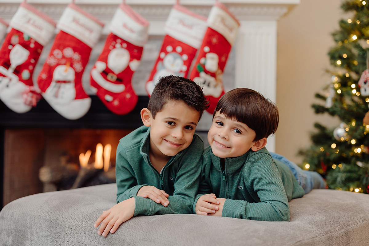 Two brothers lying on their stomachs on an ottoman in front of a fireplace decorated with Christmas stockings